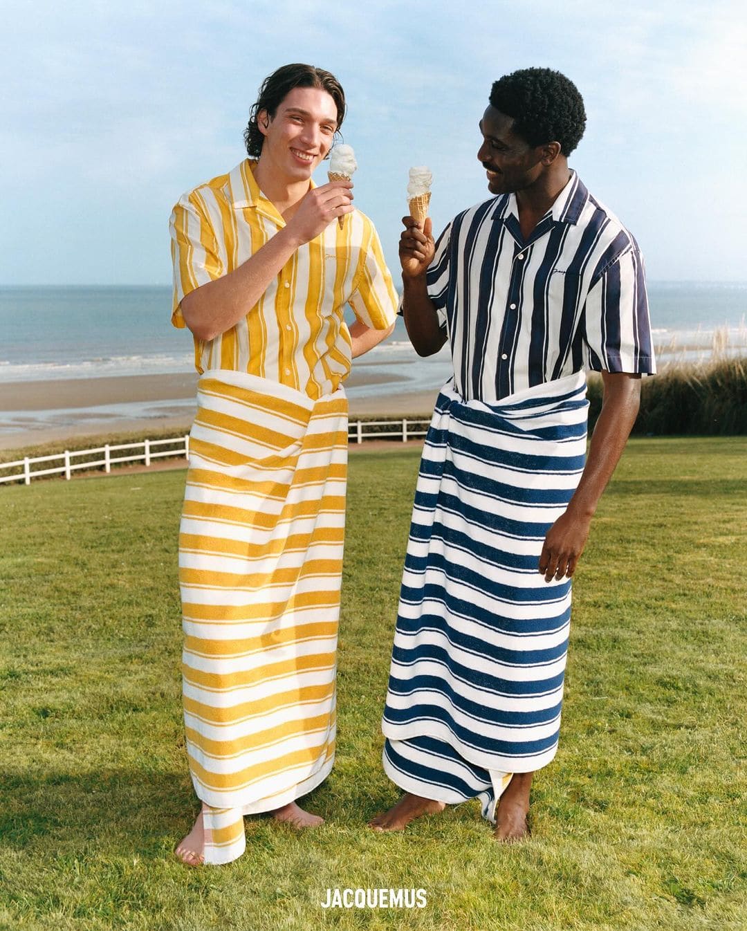 Two men on a lawn near a beach, wearing striped shirts and matching sarongs while eating ice cream cones, in a full outdoor shot.