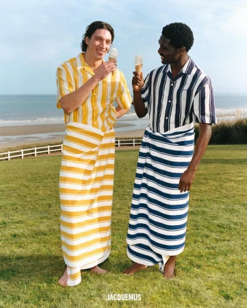 Two men on a lawn near a beach, wearing striped shirts and matching sarongs while eating ice cream cones, in a full outdoor shot.