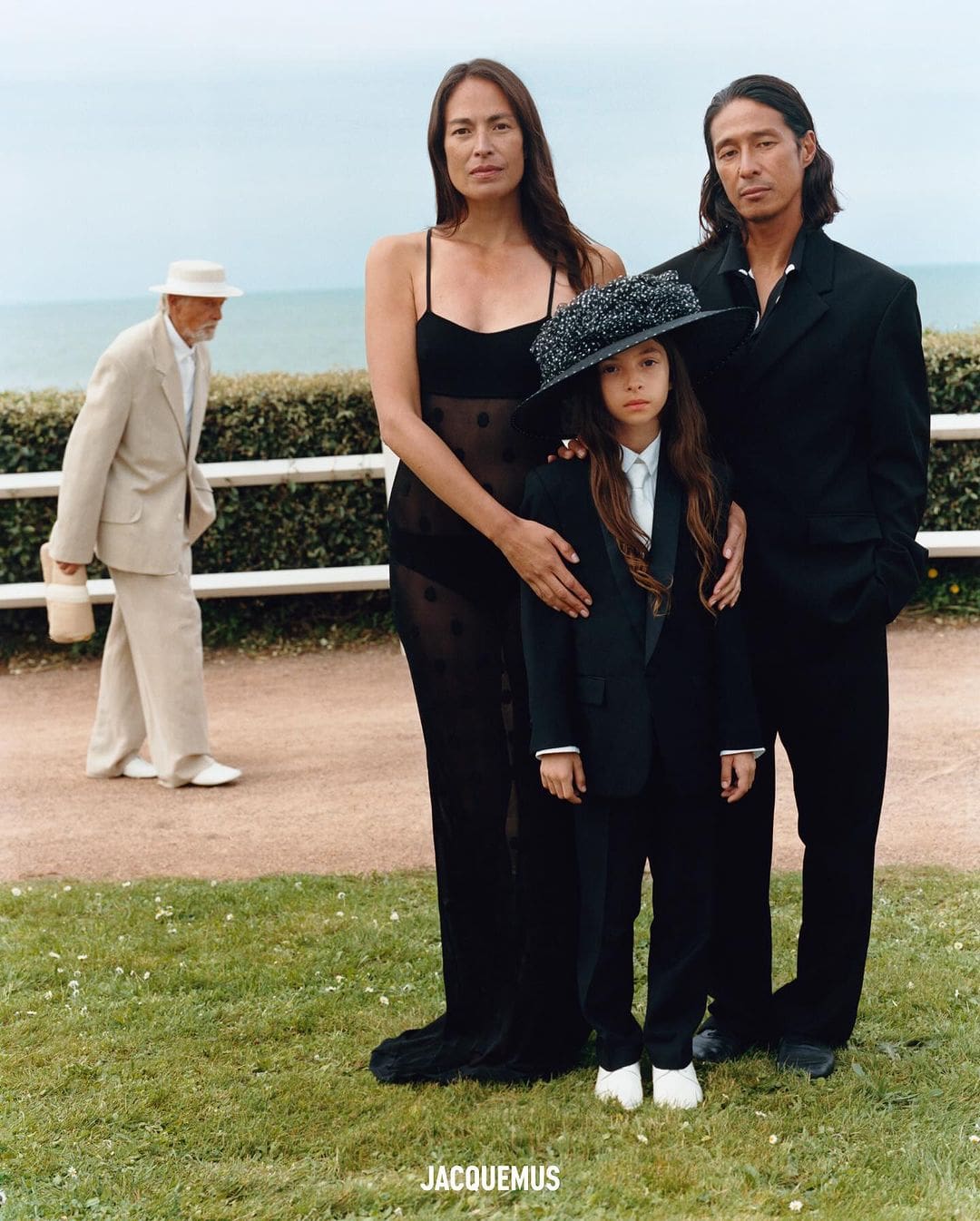 Three models posing outdoors on grass with an older man walking in the background for Jacquemus Le Mariage