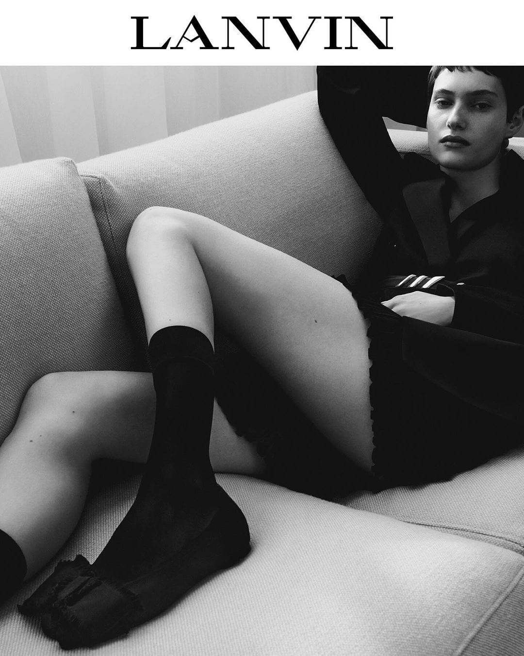 Monochrome portrait of a woman in a blazer and socks reclining on a couch, featuring the LANVIN logo at the top of the image.