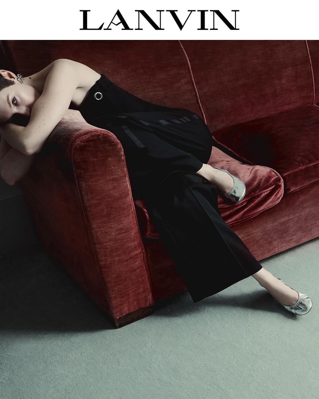 Adult female in a black dress lounges on a red velvet couch in a fashion shot; silver flats and muted lighting add to the scene.