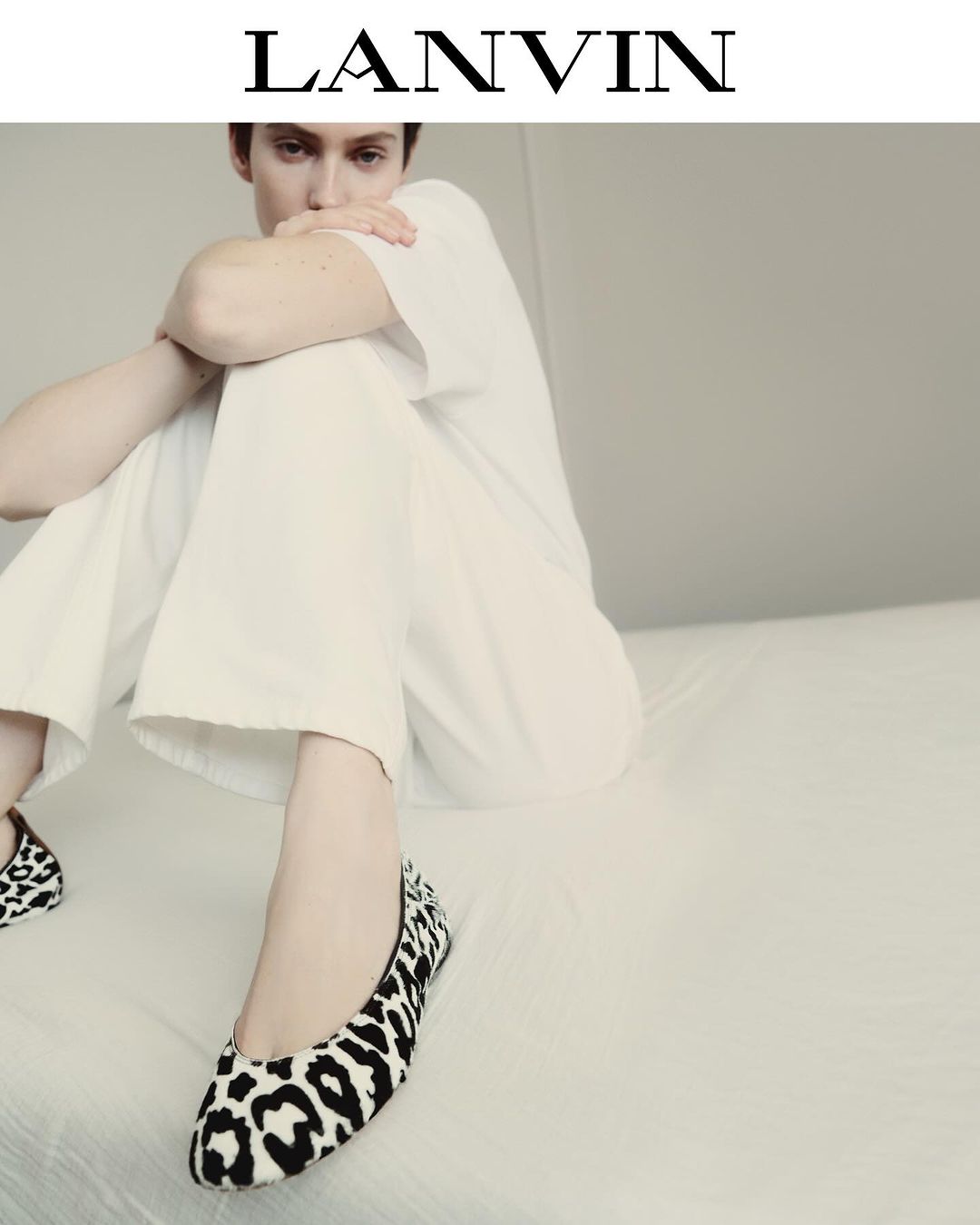 Woman in casual white outfit sits on a white bed, wearing spotted ballet flats, with Lanvin logo visible at the top of the image.