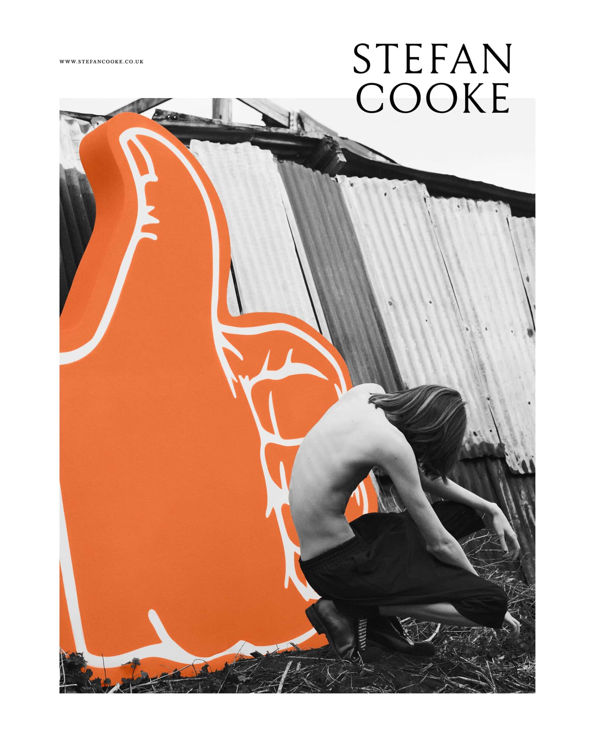 A person with long hair crouching next to a large orange foam hand in an outdoor setting, with corrugated metal in the background.