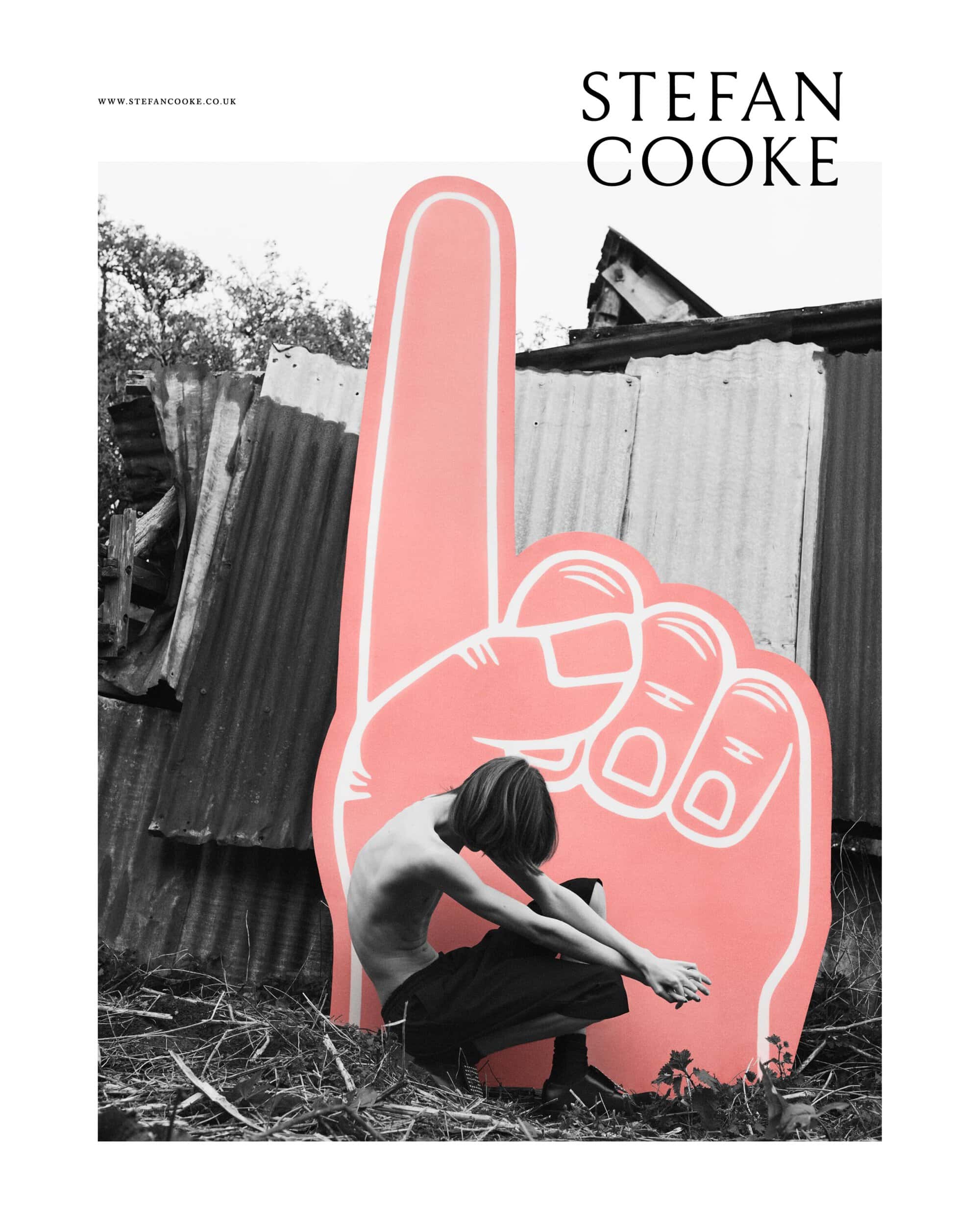 Artistic black and white portrait of a shirtless young adult crouched near a large cardboard hand outdoors, set against a corrugated fence.