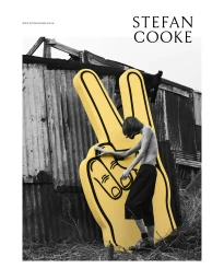 A shirtless model leaning against a large yellow hand prop in front of a rustic corrugated metal shed.