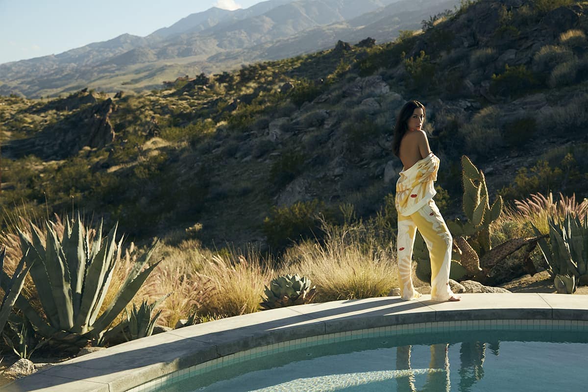 Adult female in casual patterned top and pants posing by poolside, with mountain landscape and desert flora in the background on a sunny day.