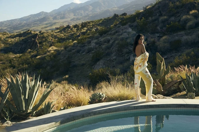 Adult female in casual patterned top and pants posing by poolside, with mountain landscape and desert flora in the background on a sunny day.
