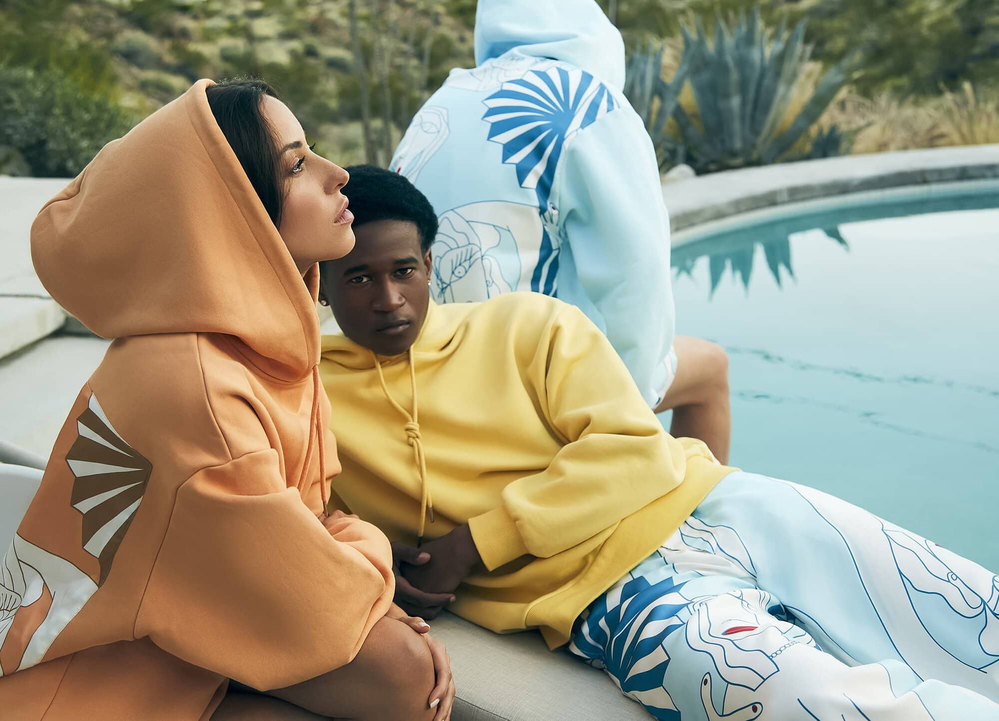 Three fashion models showcasing colourful hoodies and pants, posing poolside against a backdrop of lush greenery and architectural elements.