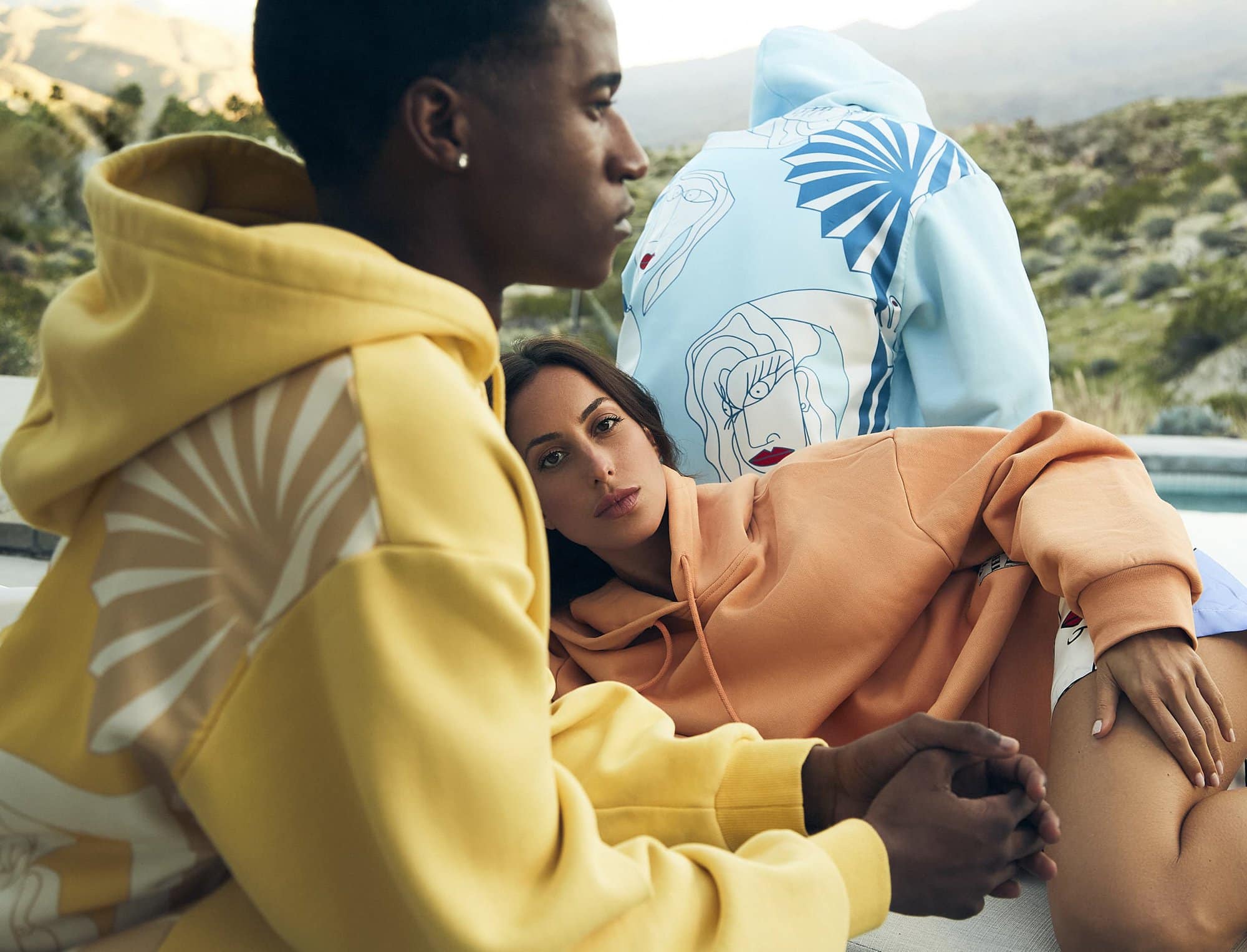 Three people in graphic hoodies pose outdoors. A brunette woman reclines in the foreground, while two others stand behind her in bright sunlight.