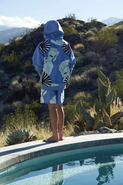 Person in blue patterned hoodie and shorts stands near a pool with a desert mountain backdrop on a sunny day.