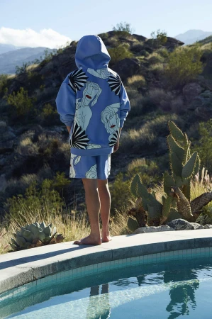Person in blue patterned hoodie and shorts stands near a pool with a desert mountain backdrop on a sunny day.