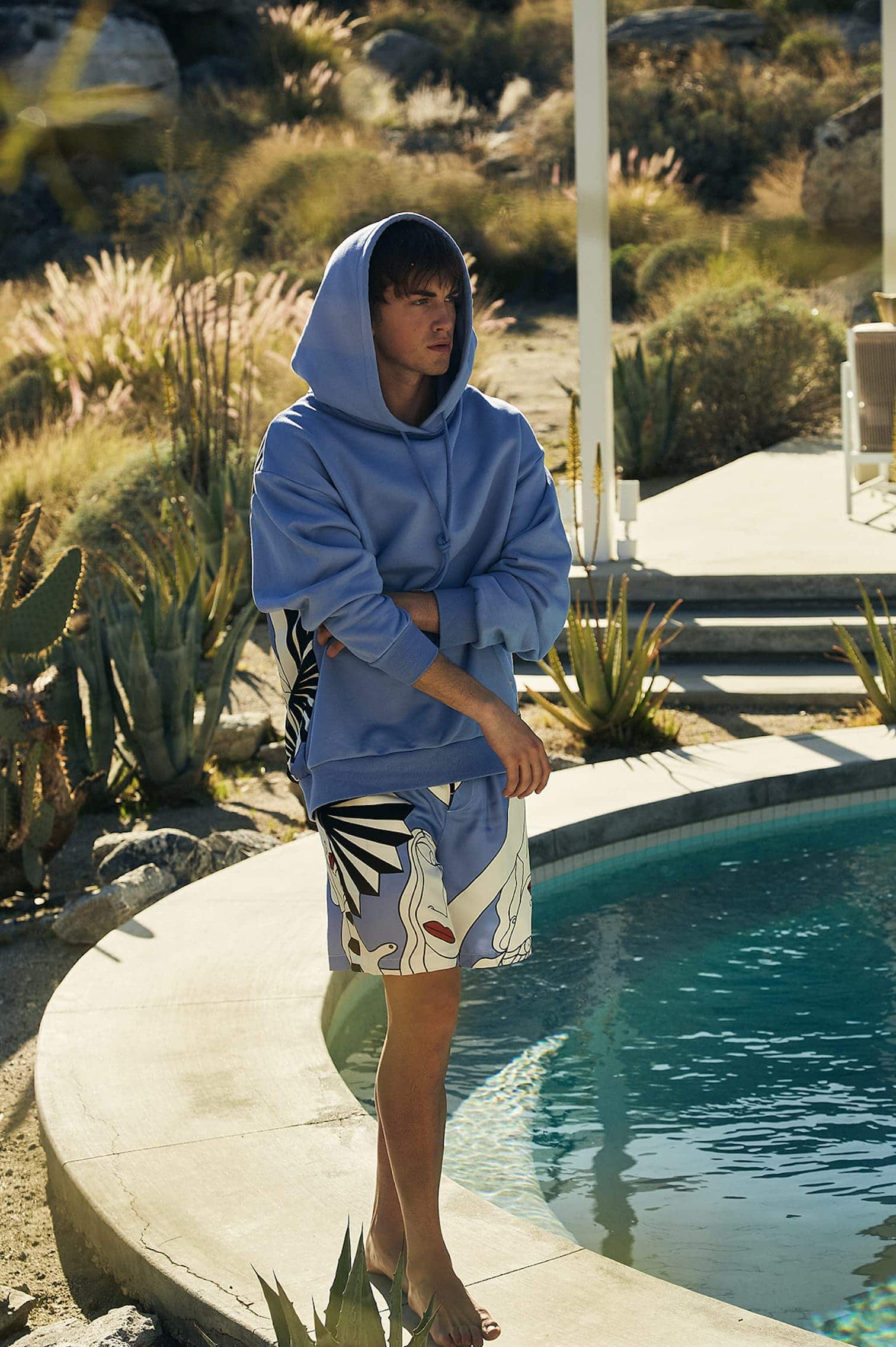 A young adult in a blue hooded sweatshirt and patterned shorts stands beside a pool outdoors in a desert landscape setting on a sunny day.