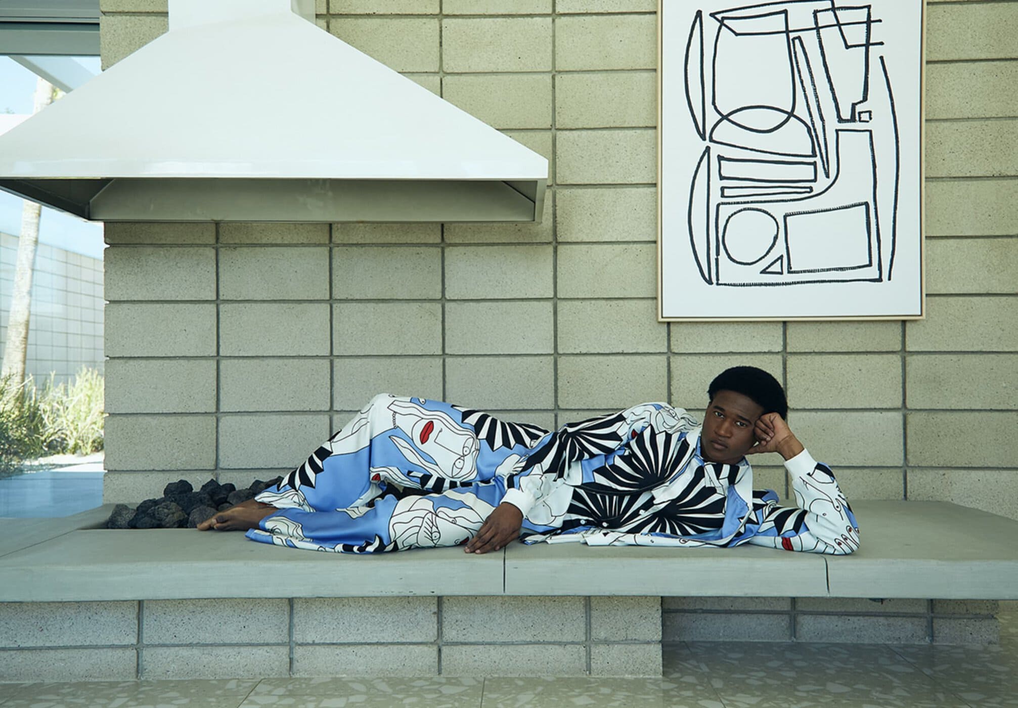 Adult male laying on a concrete surface in a patterned shirt and pants, with an abstract artwork and white range hood in the background.