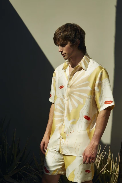 Man in patterned shirt and shorts posing outdoors in natural light with a shadow, showcasing a modern fashion style for editorial use.
