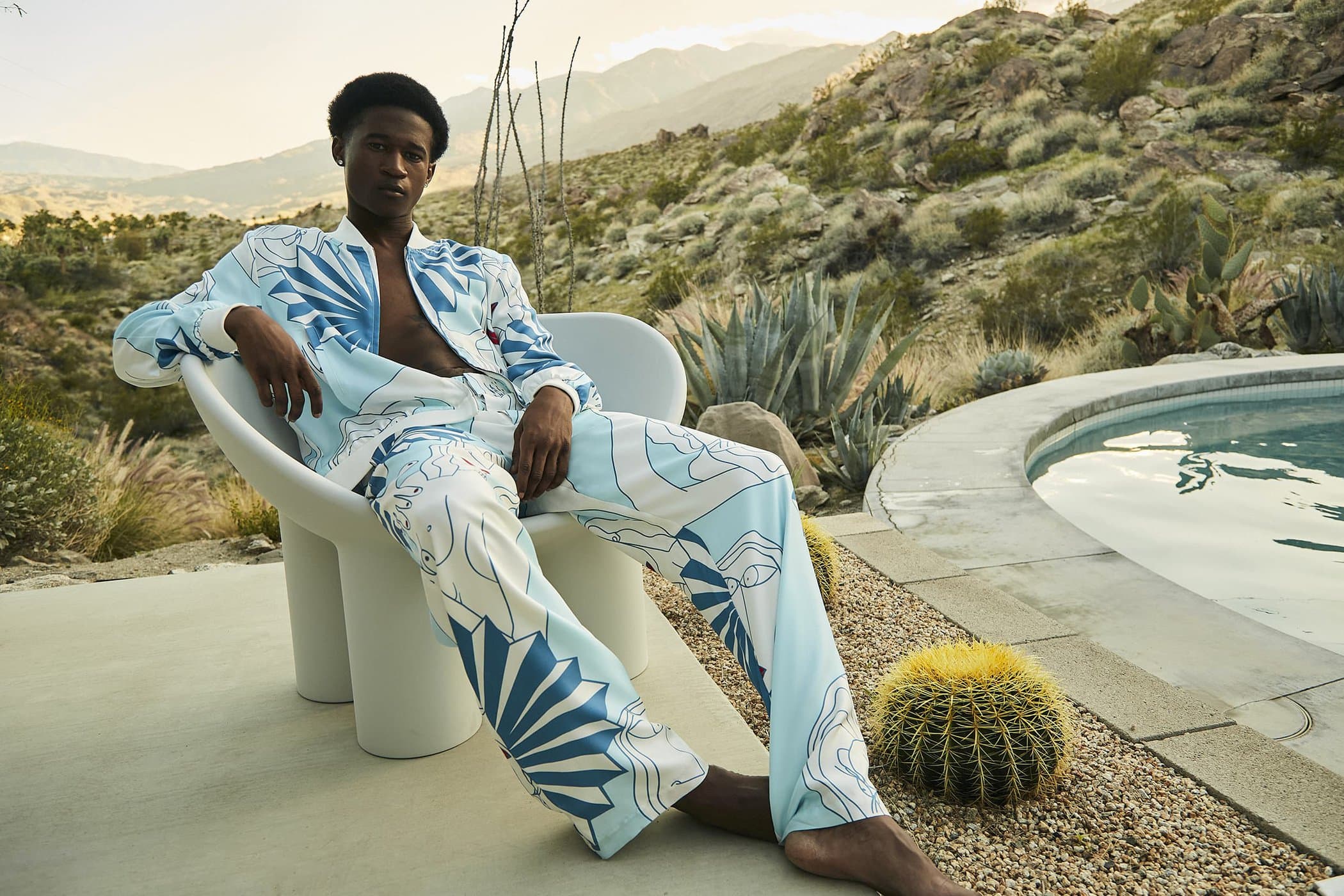 Man in patterned shirt and pants relaxes on modern chair near pool with desert mountain backdrop, capturing an outdoor fashion vibe.