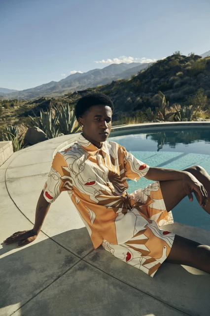 A young Black man models patterned shirt and shorts by a pool with mountain views on a sunny day for a fashion editorial.