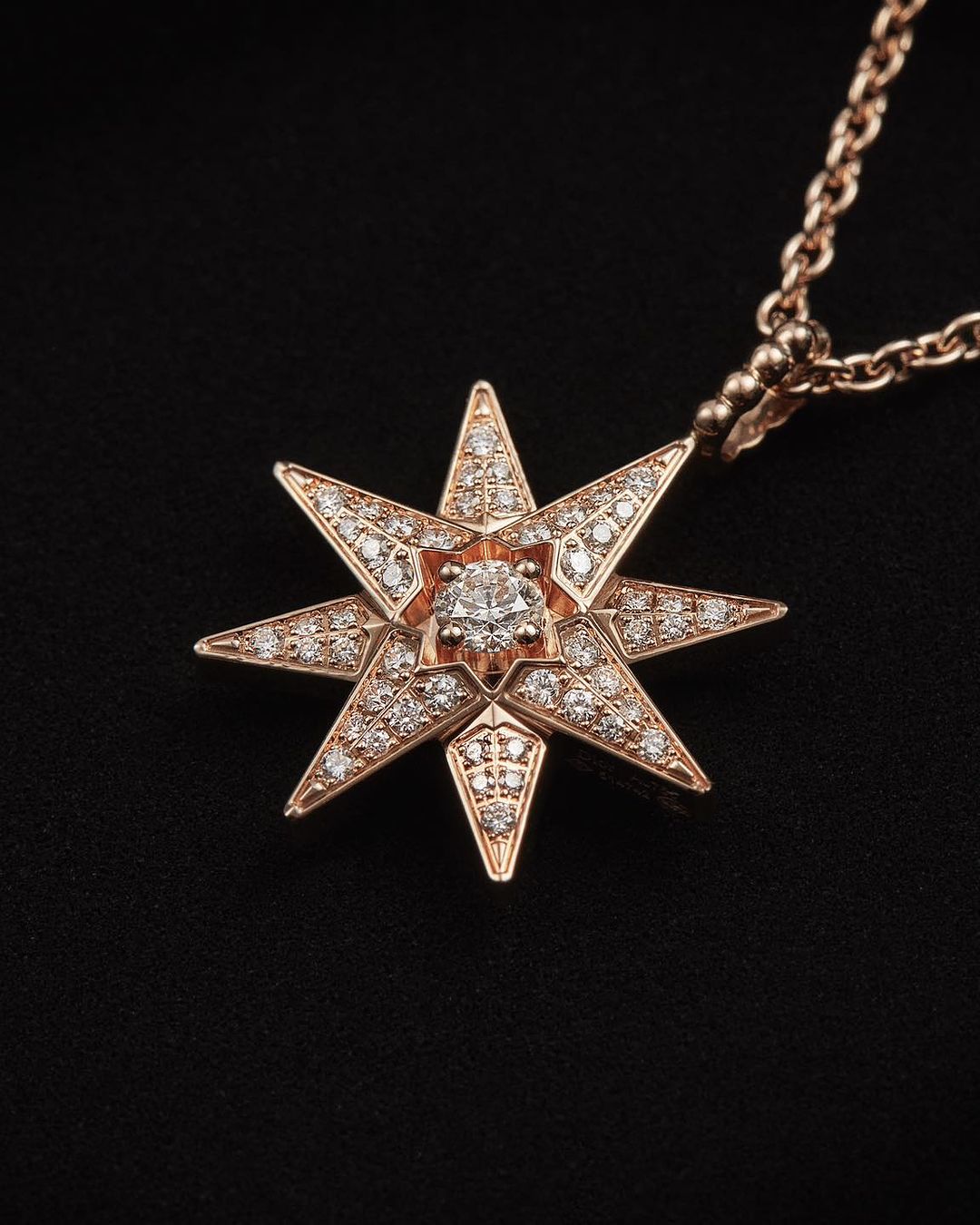 Close-up of a rose gold pendant necklace featuring a starburst design with diamonds on a black background; jewelry advertisement.
