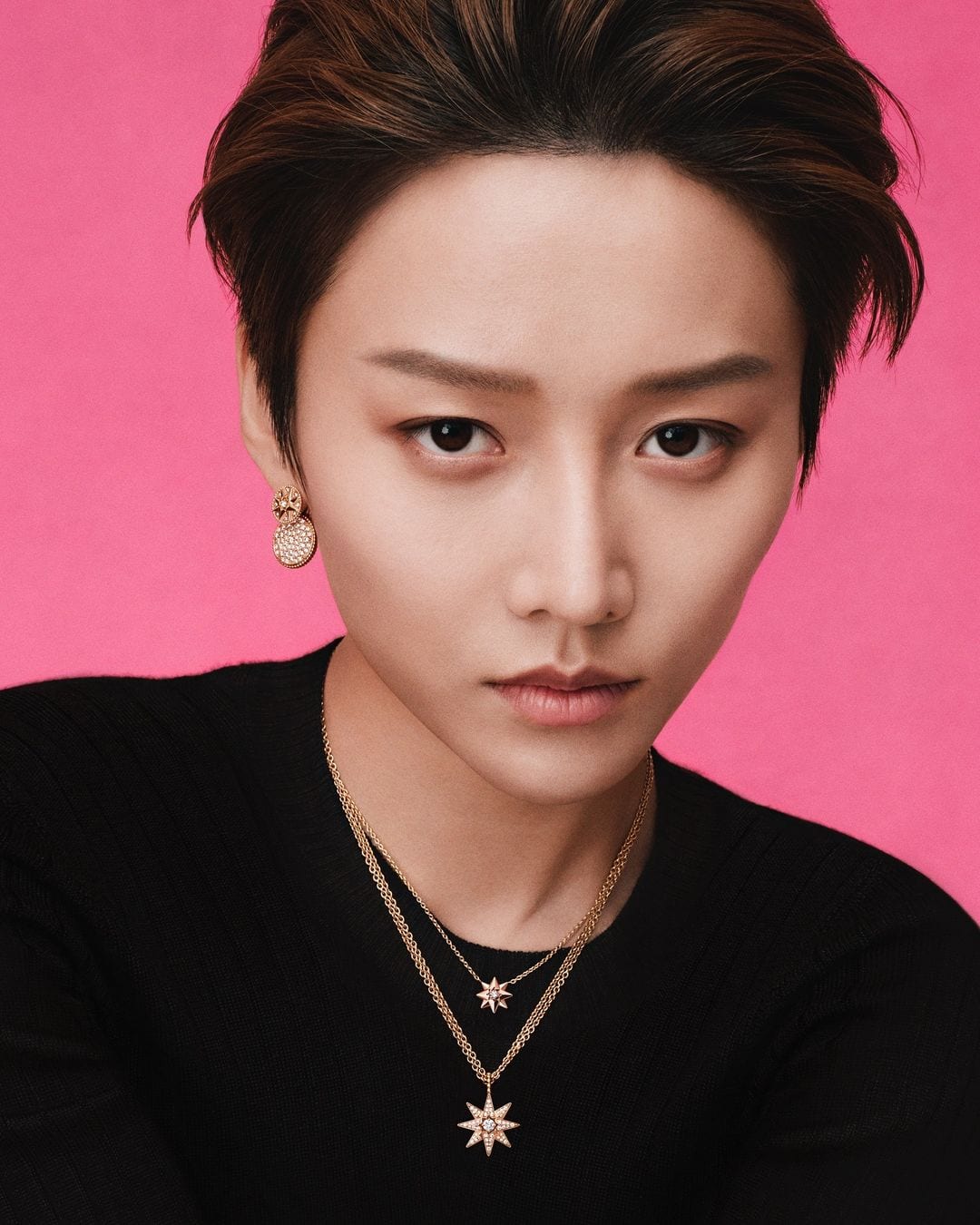 Close-up portrait of a young adult with dark hair and gold necklaces against a solid pink backdrop in a studio setting.