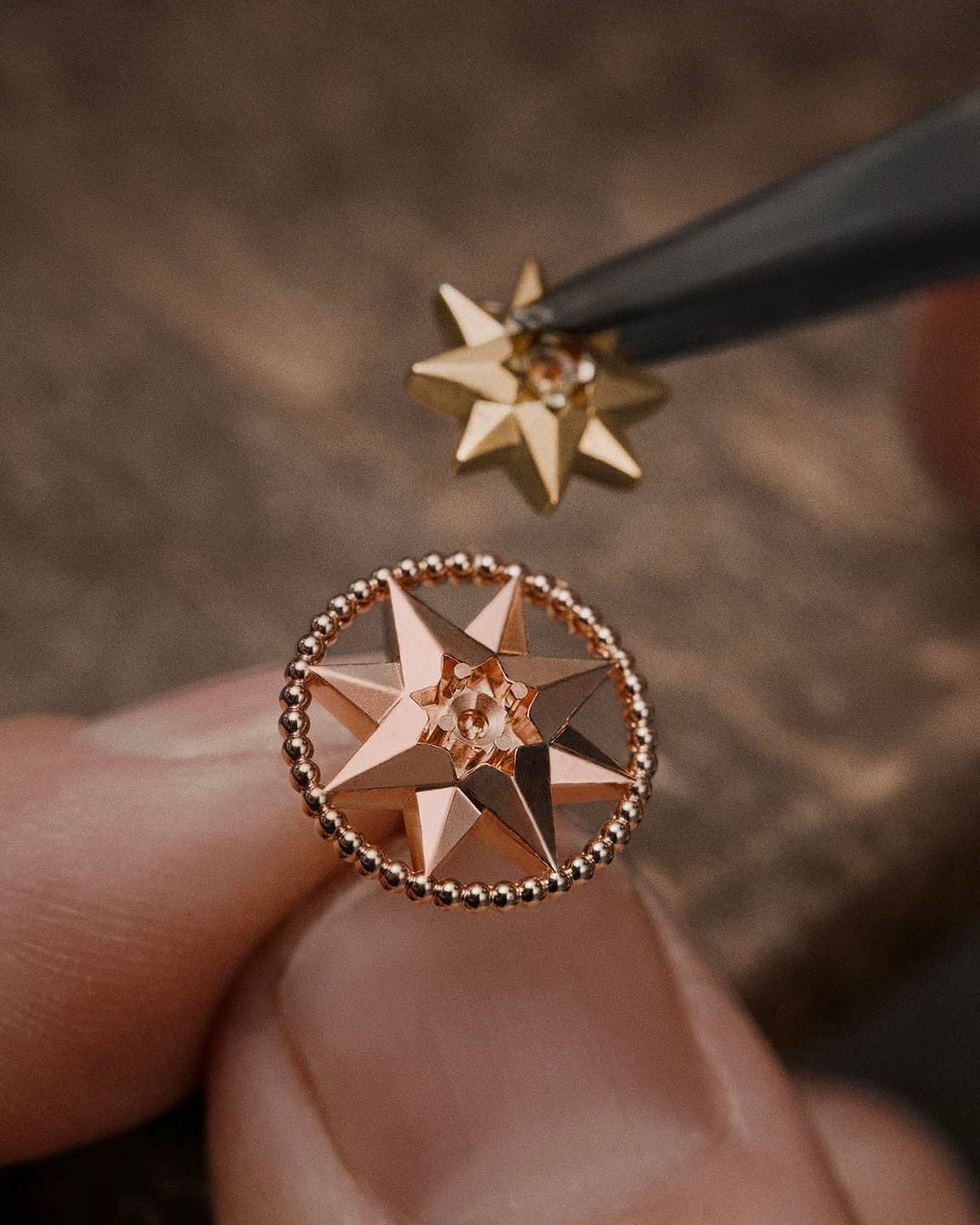 Close-up of a pair of star-shaped gold and rose-gold earrings held with tweezers against a blurred brown wooden surface.