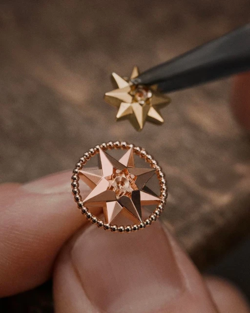 Close-up of a pair of star-shaped gold and rose-gold earrings held with tweezers against a blurred brown wooden surface.