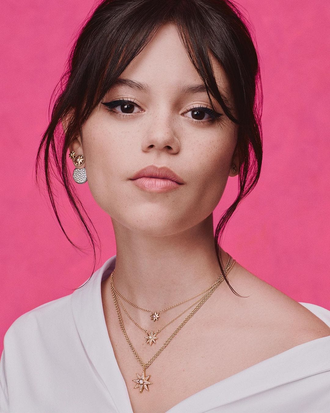 Close-up shot of a fair-skinned woman with dark hair and eye makeup, wearing gold necklaces and earrings against a pink backdrop.