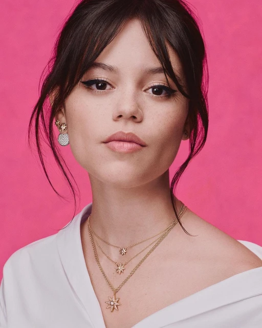 Close-up shot of a fair-skinned woman with dark hair and eye makeup, wearing gold necklaces and earrings against a pink backdrop.