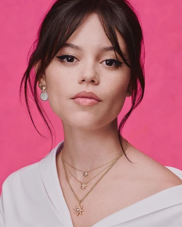 Close-up shot of a fair-skinned woman with dark hair and eye makeup, wearing gold necklaces and earrings against a pink backdrop.