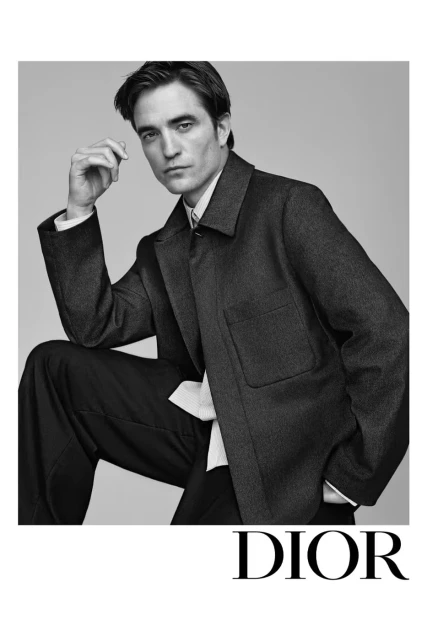 Black and white portrait of Robert Pattinson wearing a dark jacket and trousers, posing against a neutral background for a Dior advertisement.