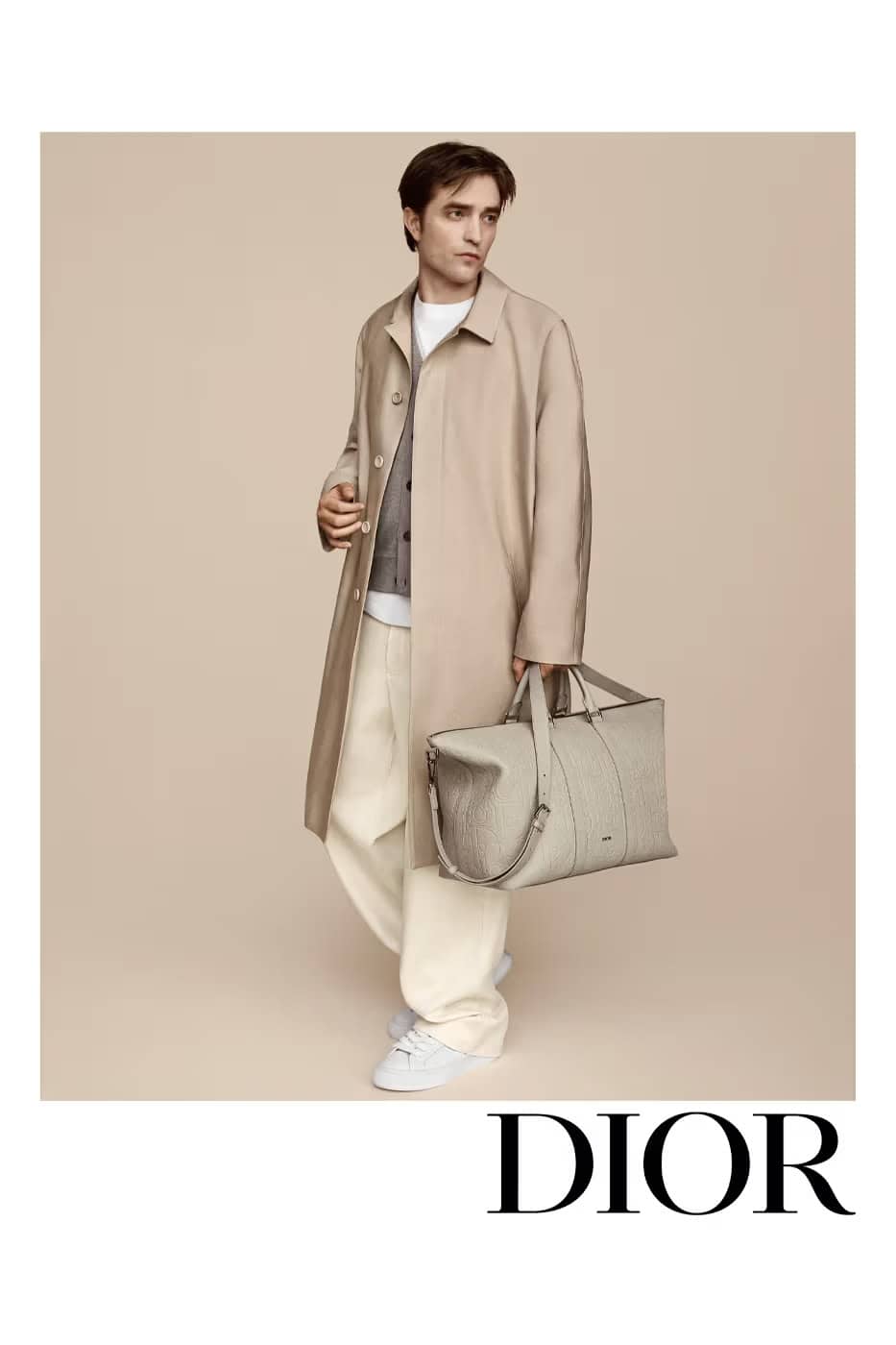 An adult male model poses in an outdoor setting wearing a beige trench coat, sweater, trousers, and white sneakers while holding a gray bag.