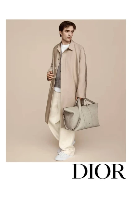 An adult male model poses in an outdoor setting wearing a beige trench coat, sweater, trousers, and white sneakers while holding a gray bag.