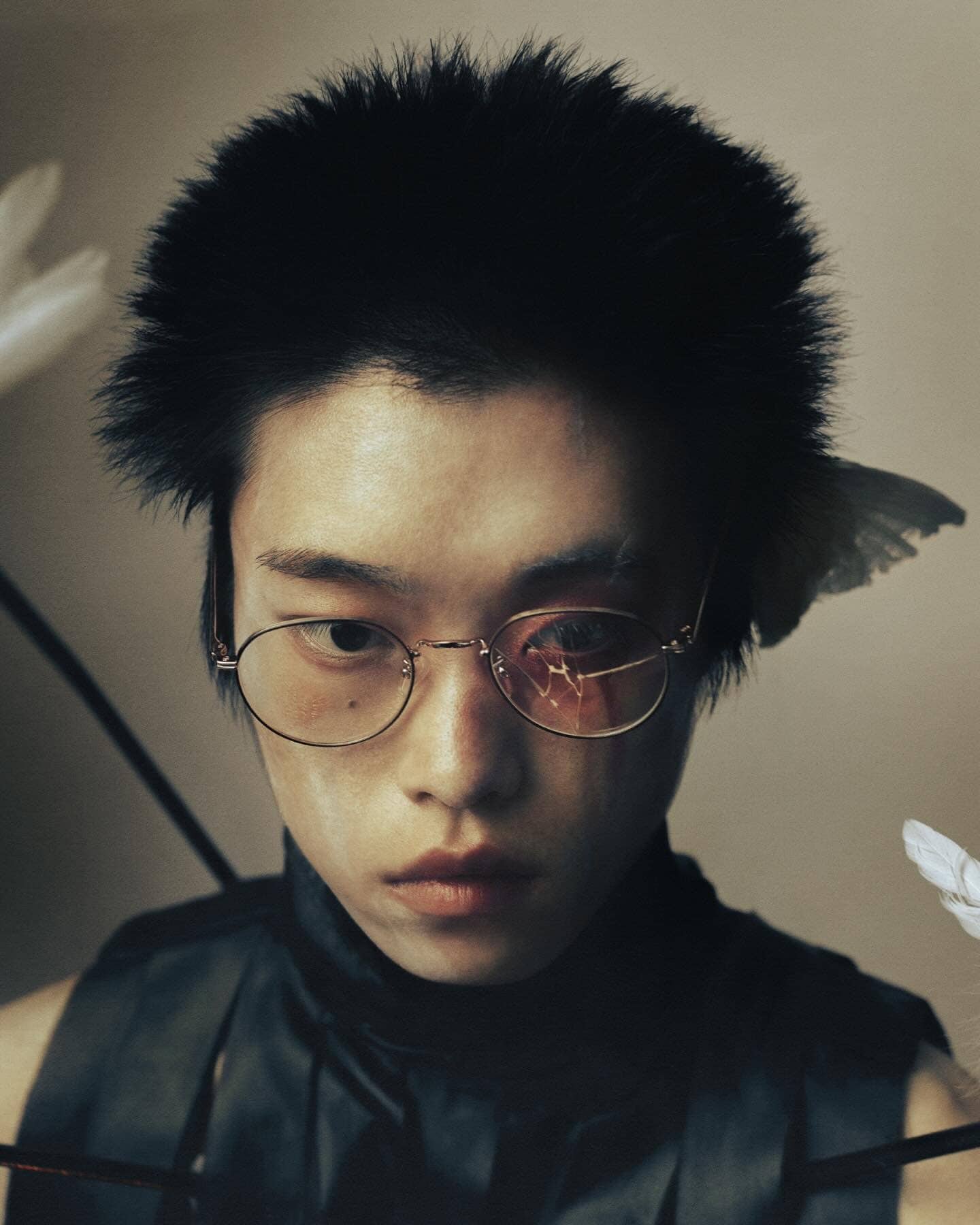 Portrait of an Asian person with arrows and feathers, wearing glasses and a black top against a neutral backdrop, creating a unique fashion statement.