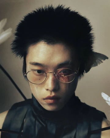 Portrait of an Asian person with arrows and feathers, wearing glasses and a black top against a neutral backdrop, creating a unique fashion statement.