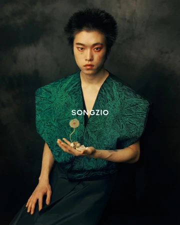 Portrait of a model with dramatic eye makeup, wearing a green textured top, holding a dried flower against a dark, moody background.