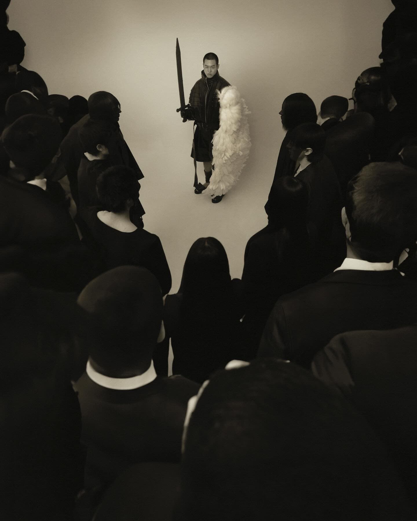 A man in a black jacket, kilt, and white feather-like adornment holds a sword, standing on a white platform, observed by a crowd in dark clothing.