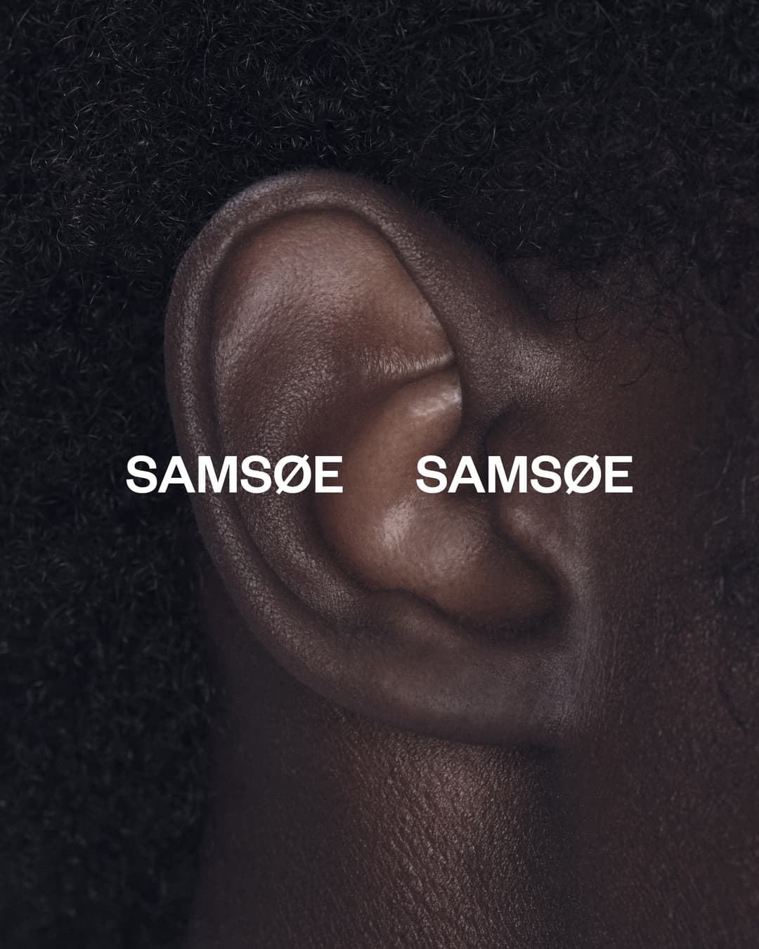 Close-up photo of human ear with 'Samsøe Samsøe' text overlaid; showcasing skin texture and black hair on a dark background.