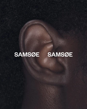 Close-up photo of human ear with 'Samsøe Samsøe' text overlaid; showcasing skin texture and black hair on a dark background.