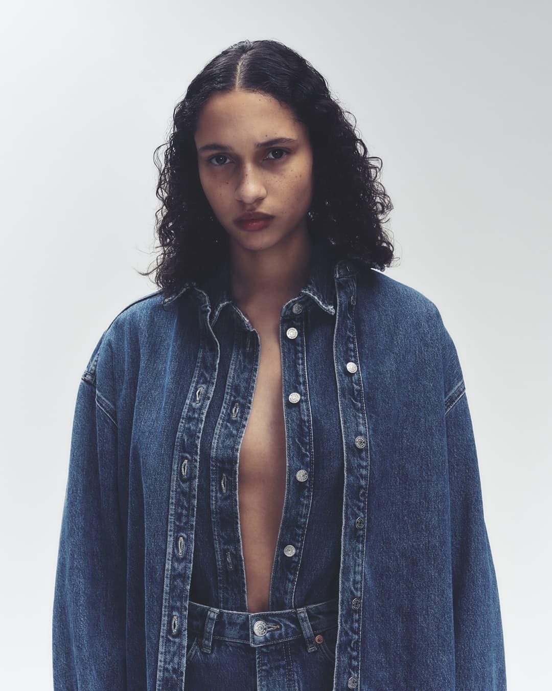 A young woman poses in an unbuttoned blue denim jacket and matching jeans against a neutral backdrop in a fashion editorial shot.