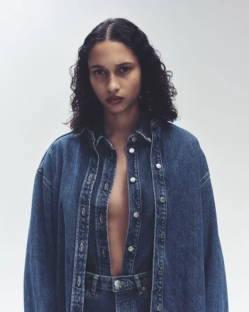 A young woman poses in an unbuttoned blue denim jacket and matching jeans against a neutral backdrop in a fashion editorial shot.