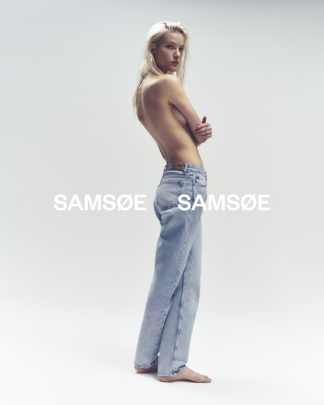 Fashion shot of a blond woman in light wash jeans, posing in a photo studio in a side view pose, with the brand logo on the backdrop.
