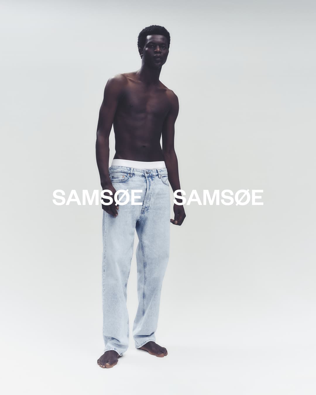 An adult Black male model poses shirtless in a studio wearing jeans, with the brand name SAMSOE visible in the bright, high-key background.