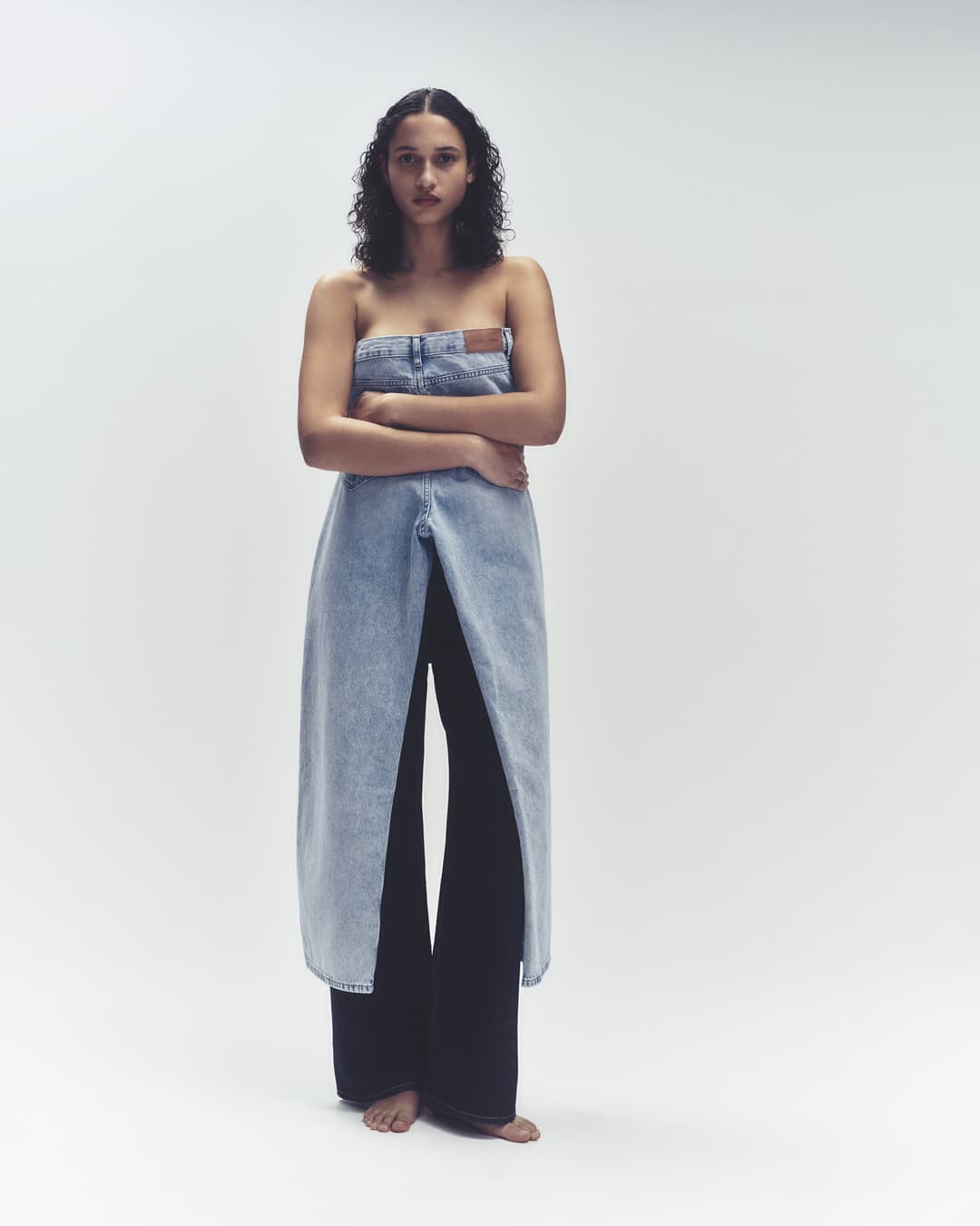 Woman in denim-inspired strapless dress and wide-leg pants stands against a bright backdrop in a fashion-forward studio portrait.