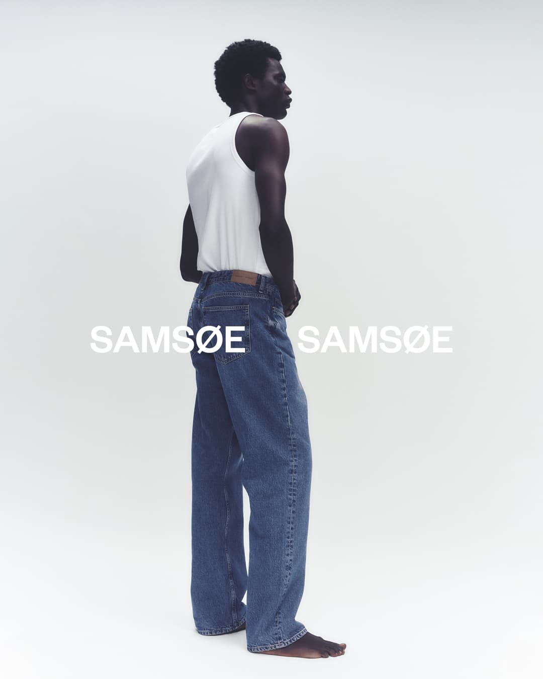 Studio shot of a Black male model wearing a white tank top and blue jeans in a fashion editorial for Samsøe Samsøe.