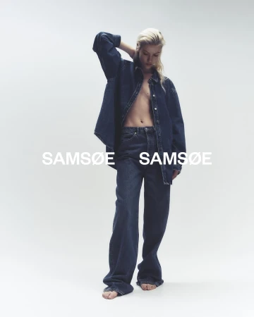A woman posing in dark-wash denim wide-leg jeans and an unbuttoned long-sleeved denim shirt on a white studio backdrop.