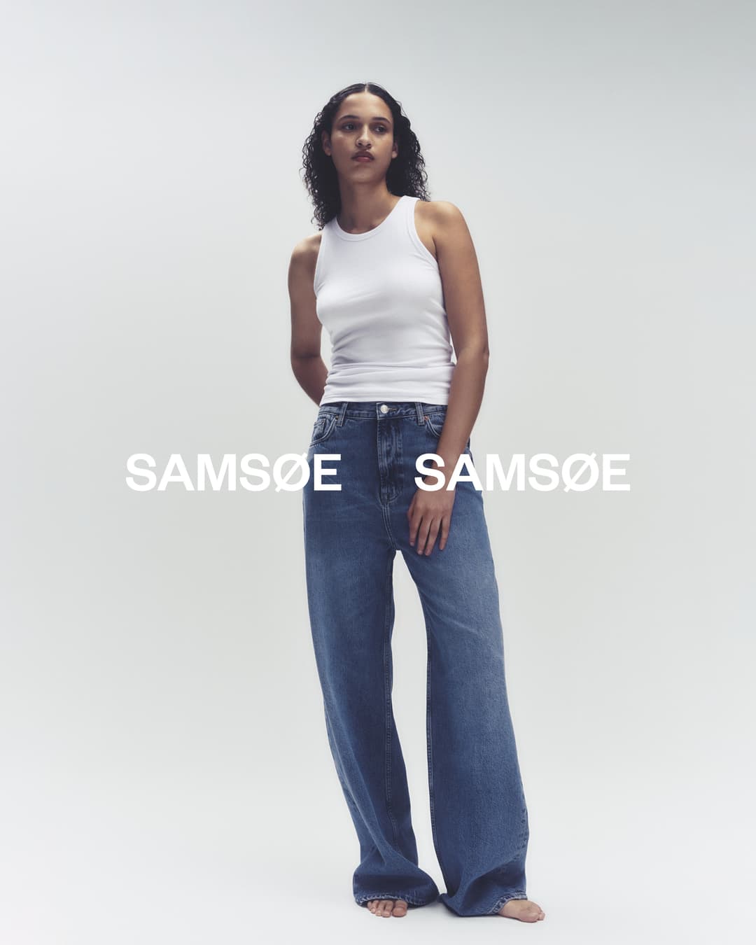 Fashion model poses in a white tank top and wide-leg blue jeans against a neutral backdrop, showcasing contemporary casual style.