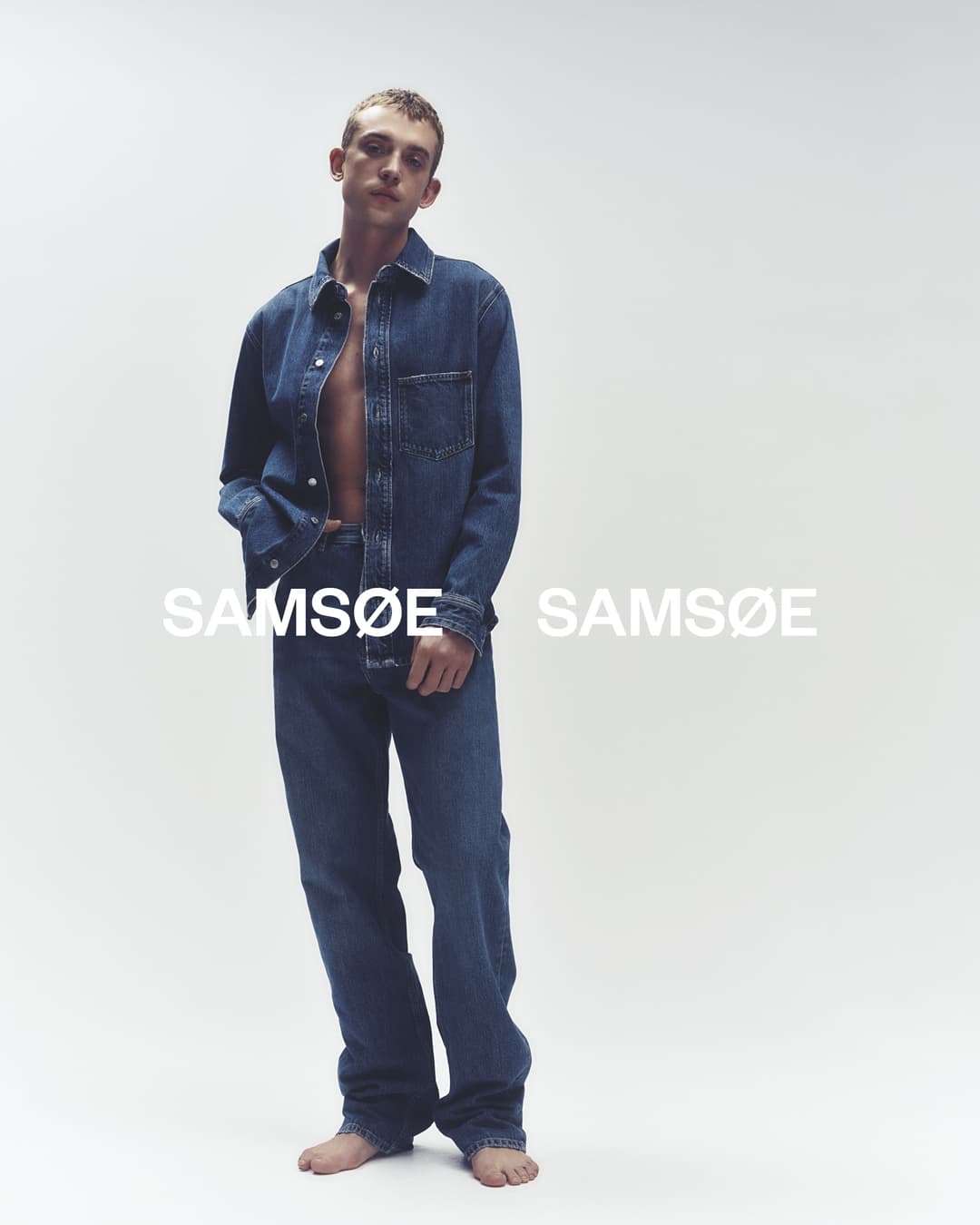 Man in denim shirt and jeans posing in a studio. Fashion shot featuring a denim outfit against a neutral backdrop with brand text.