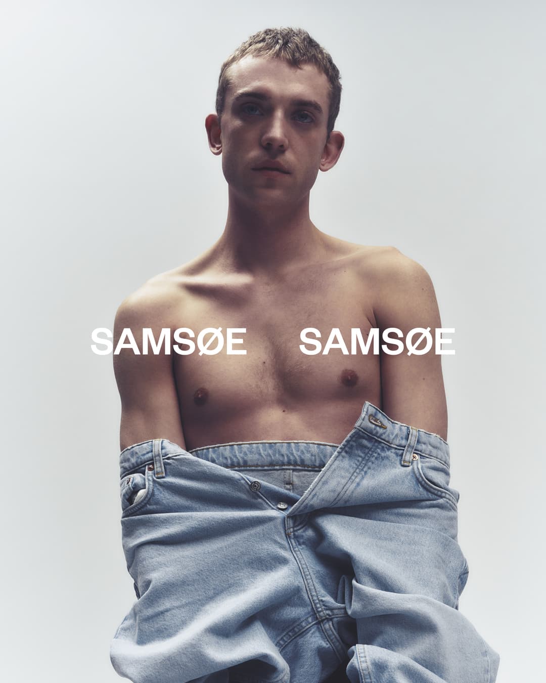 Shirtless man with jeans sitting in front of a white background. Brand logo, SAMSOE SAMSOE, is visible.