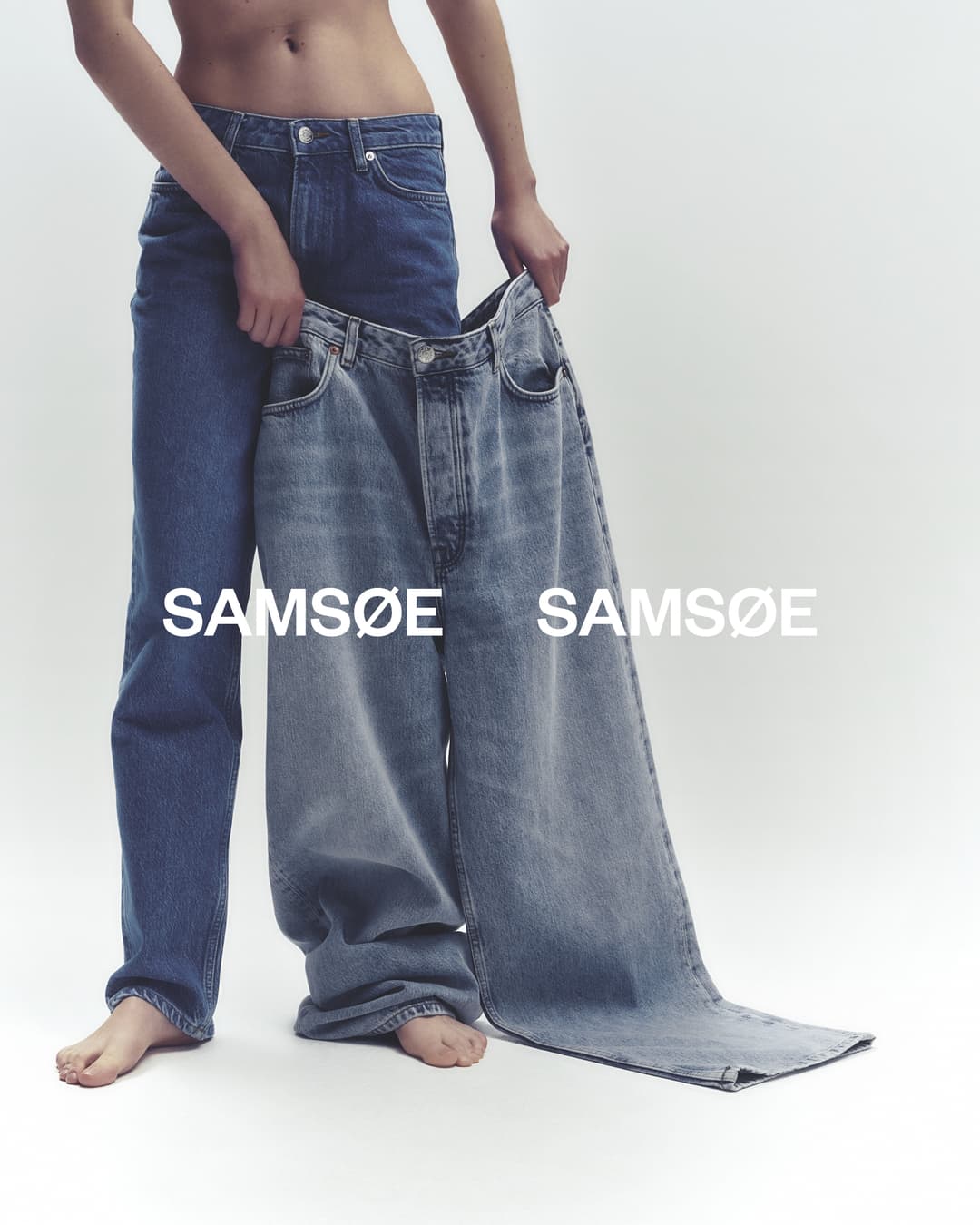 Fashionable denim jeans on a woman in a light studio. She holds a second pair of large, light-wash, wide-leg jeans in her hands.