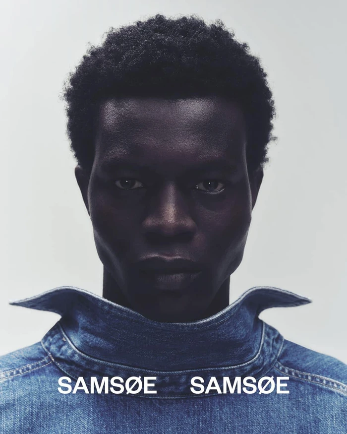Close-up portrait of a Black man with short textured hair wearing a denim jacket against a neutral grey studio background.
