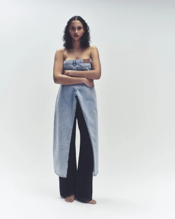 Model standing with arms crossed wearing a strapless denim top over dark trousers against a white backdrop.