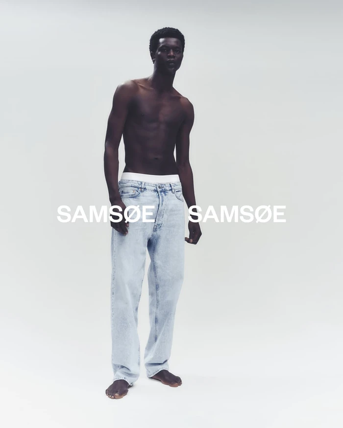 Shirtless black man wearing light wash denim jeans standing against a plain white studio background with visible branding.